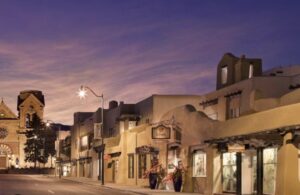 La Fonda on the Plaza at dusk seen along San Francisco Street, its stepped adobe massing and setback terraces lit against a violet sky, with the towers of the Cathedral Basilica of St. Francis further down the street.