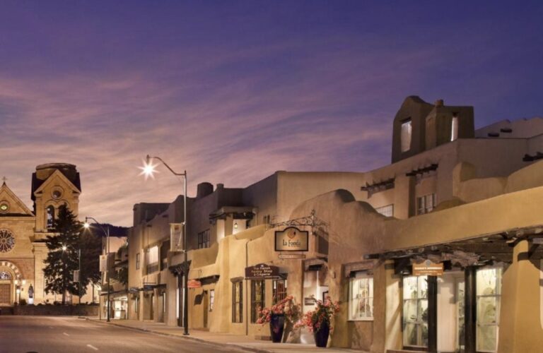 La Fonda on the Plaza at dusk seen along San Francisco Street, its stepped adobe massing and setback terraces lit against a violet sky, with the towers of the Cathedral Basilica of St. Francis further down the street.