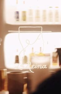 The La Reina sign behind the bar, a single line drawing of a face beneath a wide brimmed hat with the name in script below it, glowing against shelves of bottles.