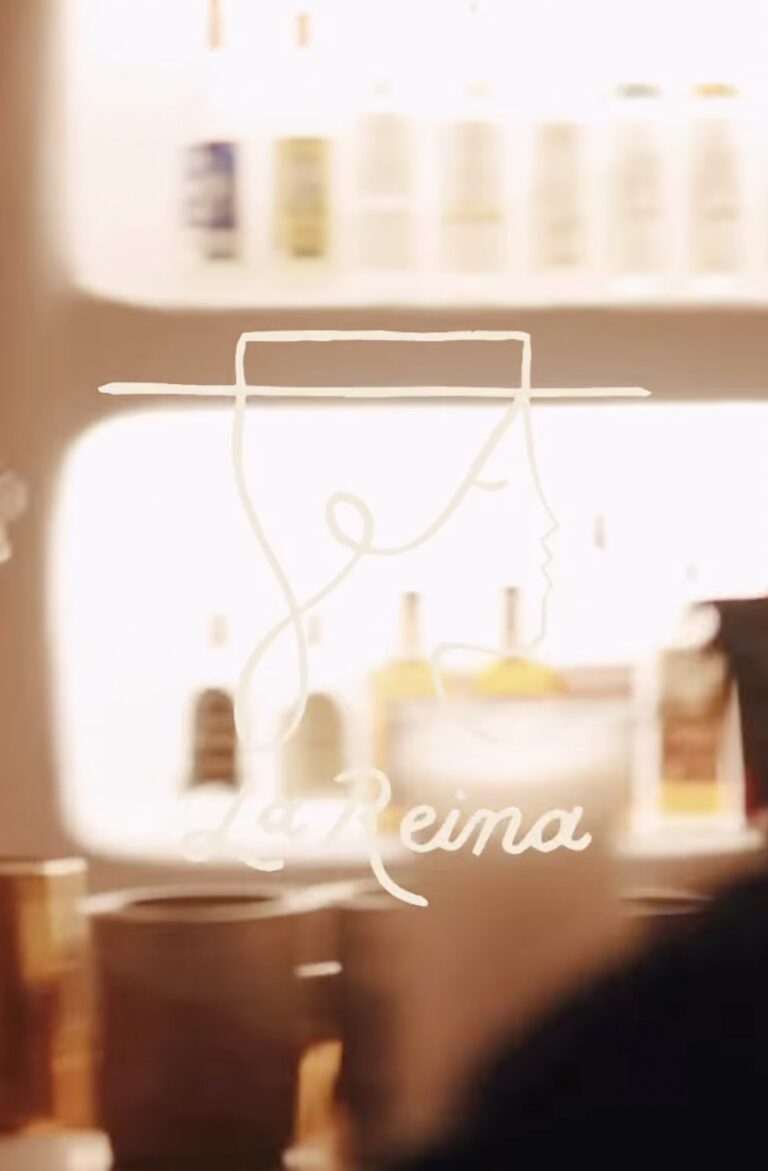 The La Reina sign behind the bar, a single line drawing of a face beneath a wide brimmed hat with the name in script below it, glowing against shelves of bottles.