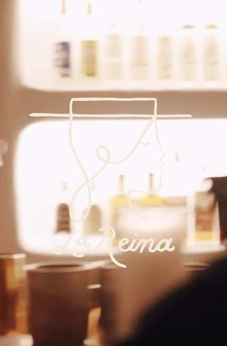 The La Reina sign behind the bar, a single line drawing of a face beneath a wide brimmed hat with the name in script below it, glowing against shelves of bottles.