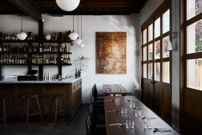 The interior of Leo's in Santa Fe: a marble-topped bar backed by shelves of bottles, walnut stools and long communal tables set with glasses and spoons, white painted block walls under an exposed wood ceiling, and tall multi-pane doors along one side where the building's garage doors used to be.