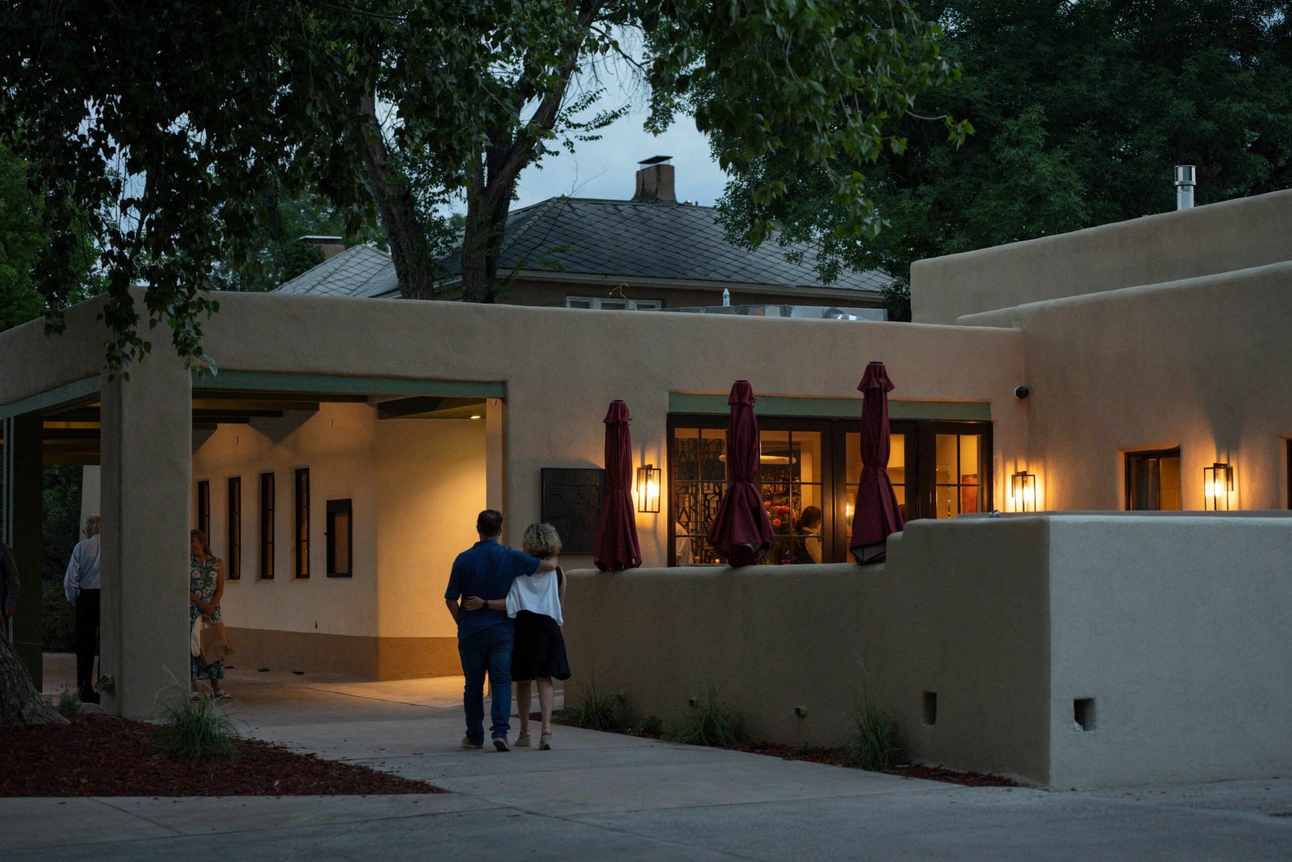 Market Steer Steakhouse at dusk on Washington Avenue in Santa Fe: a low stuccoed building with warm light in its windows, closed burgundy patio umbrellas along a low wall, lanterns lit on the facade, a steer sign beside the entrance, and two guests walking in past cottonwoods.