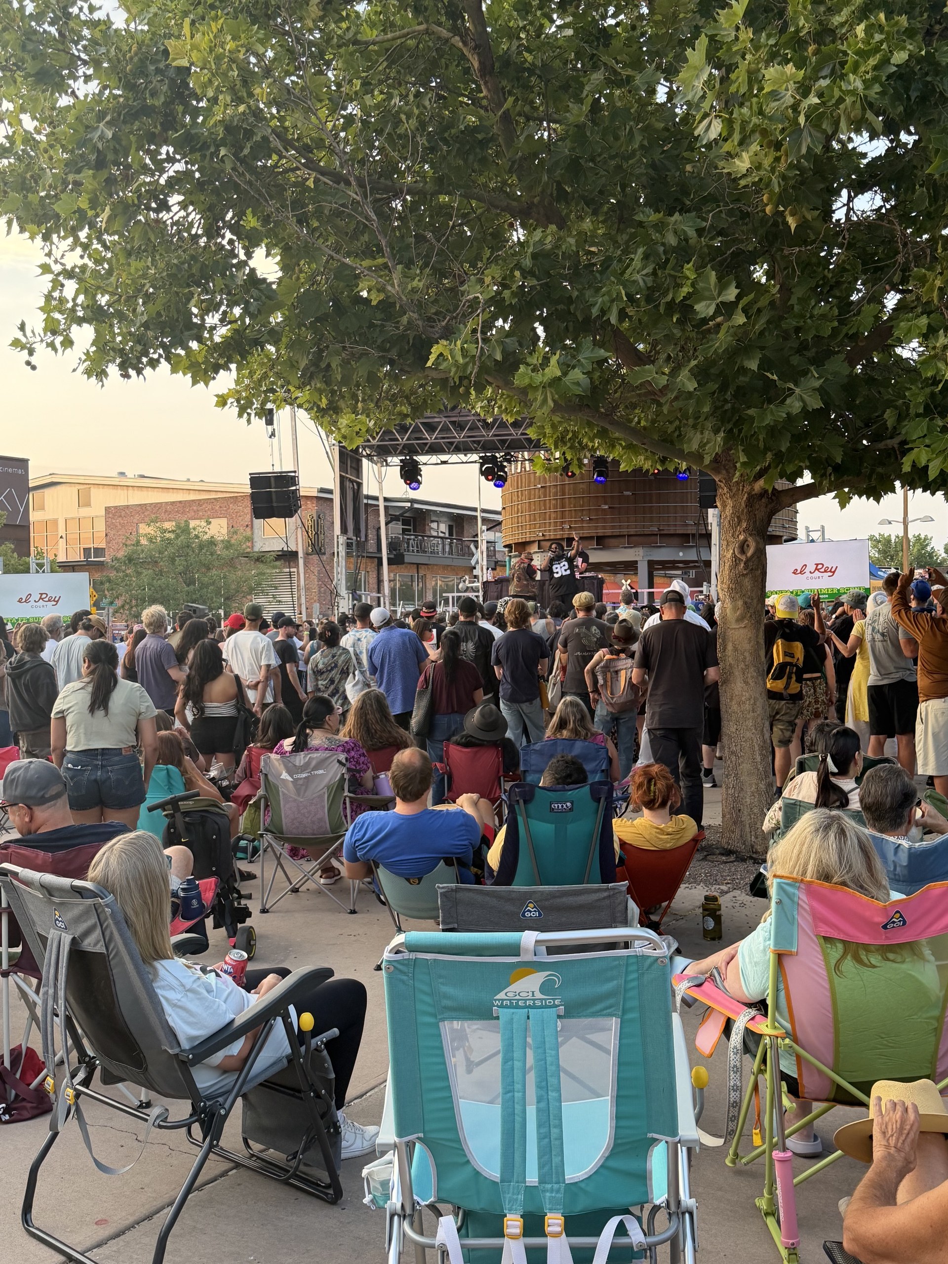 A multigenerational crowd in lawn chairs and strollers under the trees at the Santa Fe Railyard Plaza during the Hip Hop Festival, the stage and water tower beyond, August 2026.
