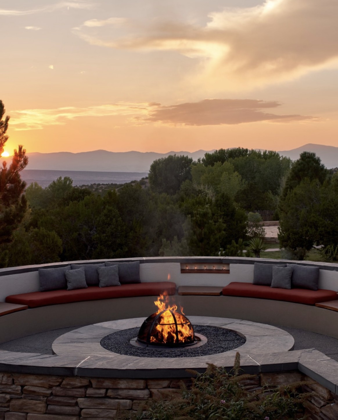 A round stone fire pit burning on a terrace at Rancho Encantado, curved banquette seating seat around it, with pinon hills and distant mountains beyond at sunset.
