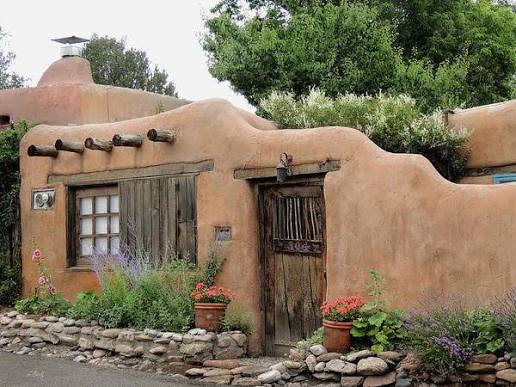 A mud plastered adobe wall along a Santa Fe lane, presenting the street a plain face: a weathered wooden plank gate set into the wall, a single small deep set window with divided panes and an old wooden lintel, projecting viga ends along the parapet above, a chimney with a metal cap at the left, hollyhocks and lavender growing at the base, potted geraniums beside the gate, a low stone retaining course along the sidewalk, and trees and shrubs showing over the top of the wall from the courtyard behind it.