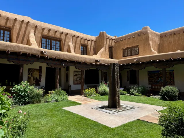 A Santa Fe placita, or enclosed courtyard, wrapped on all sides by a portal: peeled wooden posts and carved corbels carrying a beamed roof, projecting viga ends and canales along the adobe parapet above, a two story adobe elevation with deep set multi pane windows, painted and carved panels hung under the portal, and a clipped lawn with flower beds around a paved center and a standing timber post.