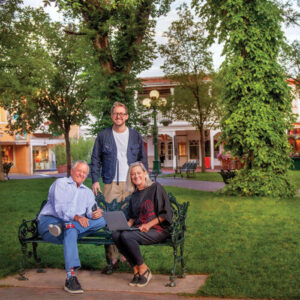 Three people on a wrought-iron bench beneath the trees on the Santa Fe Plaza, with the portal storefronts and a street lamp behind them.