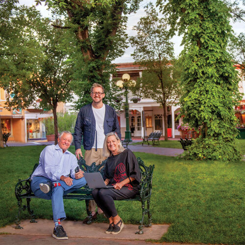 Three people on a wrought-iron bench beneath the trees on the Santa Fe Plaza, with the portal storefronts and a street lamp behind them.