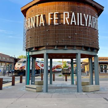 The restored wooden water tower lettered SANTA FE RAILYARD, standing on steel legs over a paved plaza in the Railyard district, with a Rail Runner train stopped on the track behind it and low brick and adobe buildings along the far side.