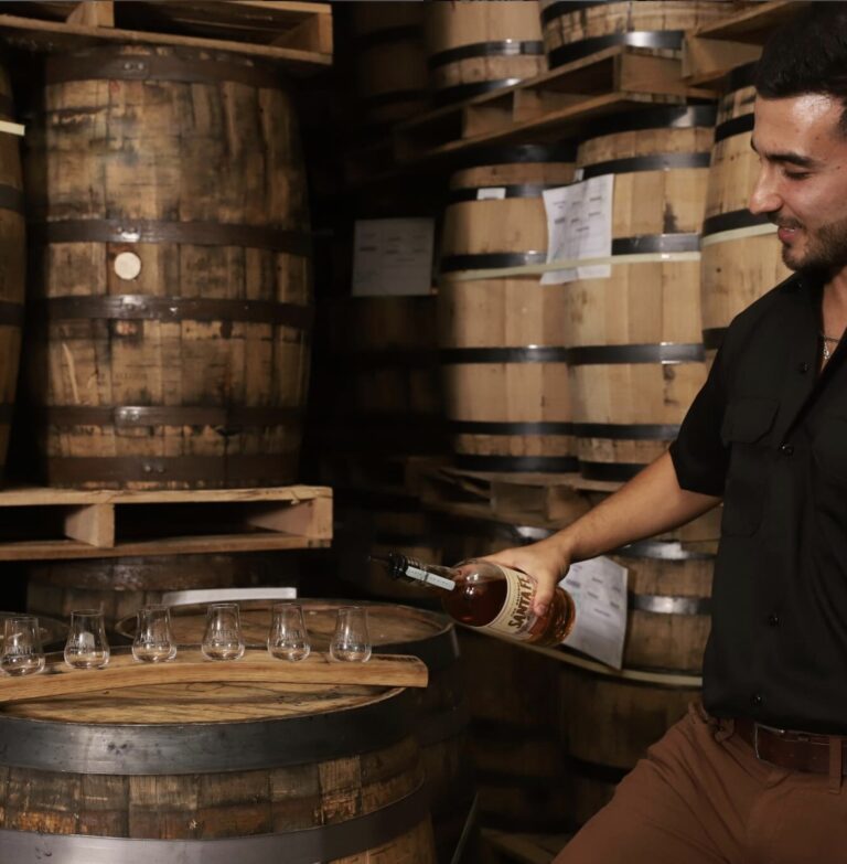A member of staff in a Santa Fe Spirits shirt pours whiskey into a row of tasting glasses set out on a barrel head, in a warehouse stacked with oak barrels on pallets.