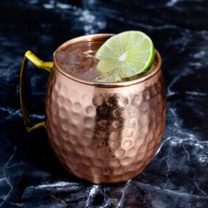 A Moscow Mule in a hammered copper mug, garnished with a wheel of lime and beaded with condensation, standing on a dark marble surface.