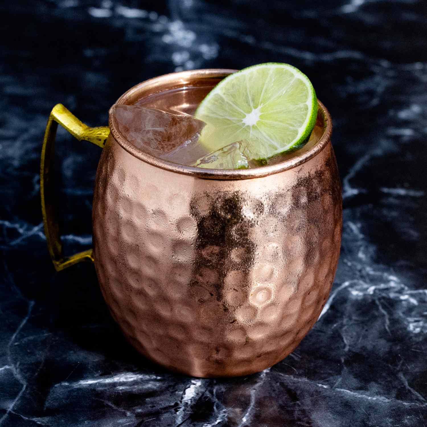 A Moscow Mule in a hammered copper mug, garnished with a wheel of lime and beaded with condensation, standing on a dark marble surface.