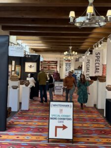 Attendees at the 48th Whitehawk Antique Indian and Ethnographic Art Show, with a Special Exhibit sign, dealer booths, Navajo textiles and pottery, Santa Fe, August 2026.