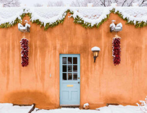 A snow covered adobe wall in Santa Fe, ristras hanging either side of a pale blue door beneath an evergreen garland