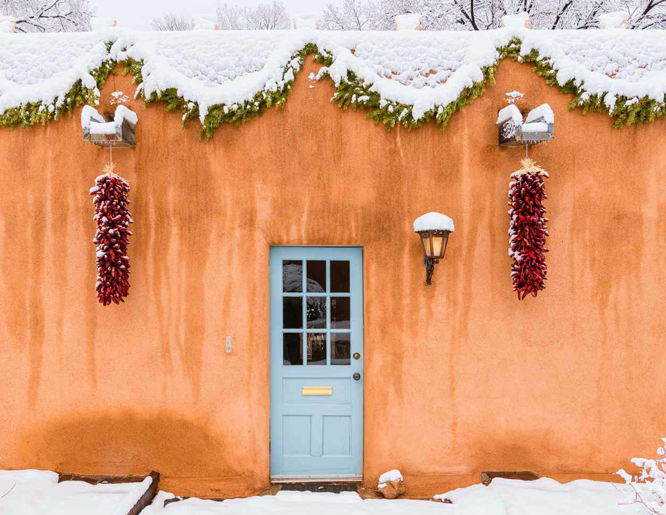 A snow covered adobe wall in Santa Fe, ristras hanging either side of a pale blue door beneath an evergreen garland