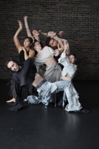 Dancers of Zeitgeist Dance Theatre in a sculptural, intertwined group pose against a brick wall, in flowing black, cream, and distressed-denim costumes.