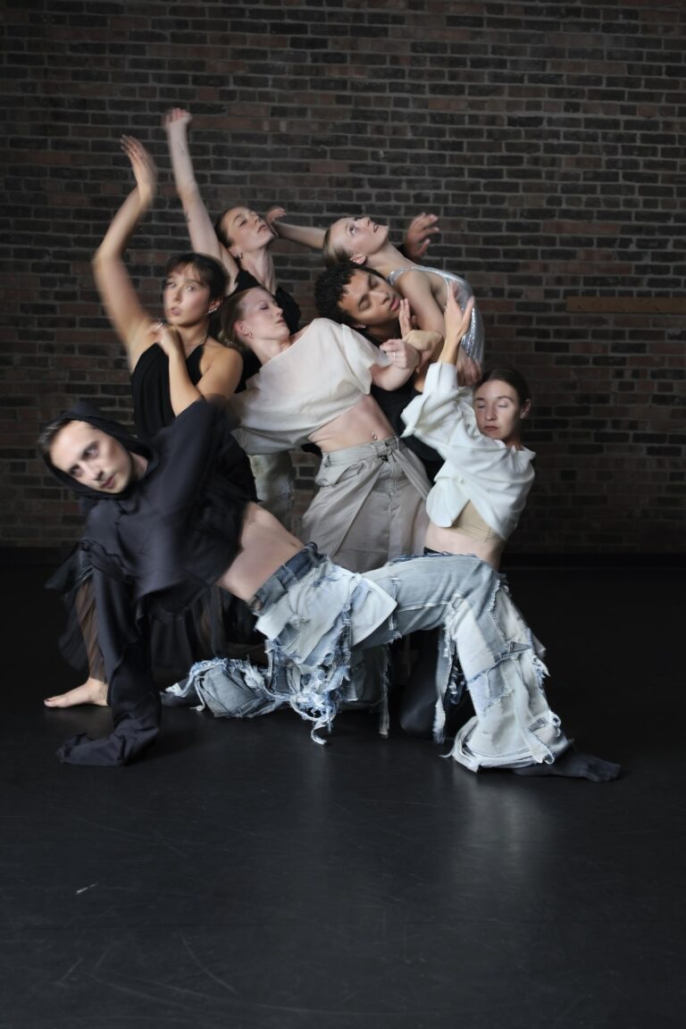 Dancers of Zeitgeist Dance Theatre in a sculptural, intertwined group pose against a brick wall, in flowing black, cream, and distressed-denim costumes.