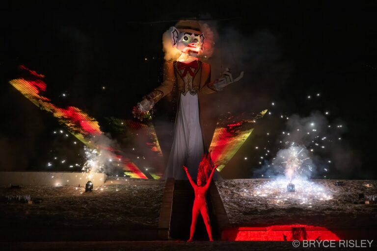 Zozobra, the fifty-foot marionette known as Old Man Gloom, alight at the head with arms outstretched over Fort Marcy Park at night, as the Fire Spirit in red raises both arms at the base of the platform and fireworks and sparks burst on either side.