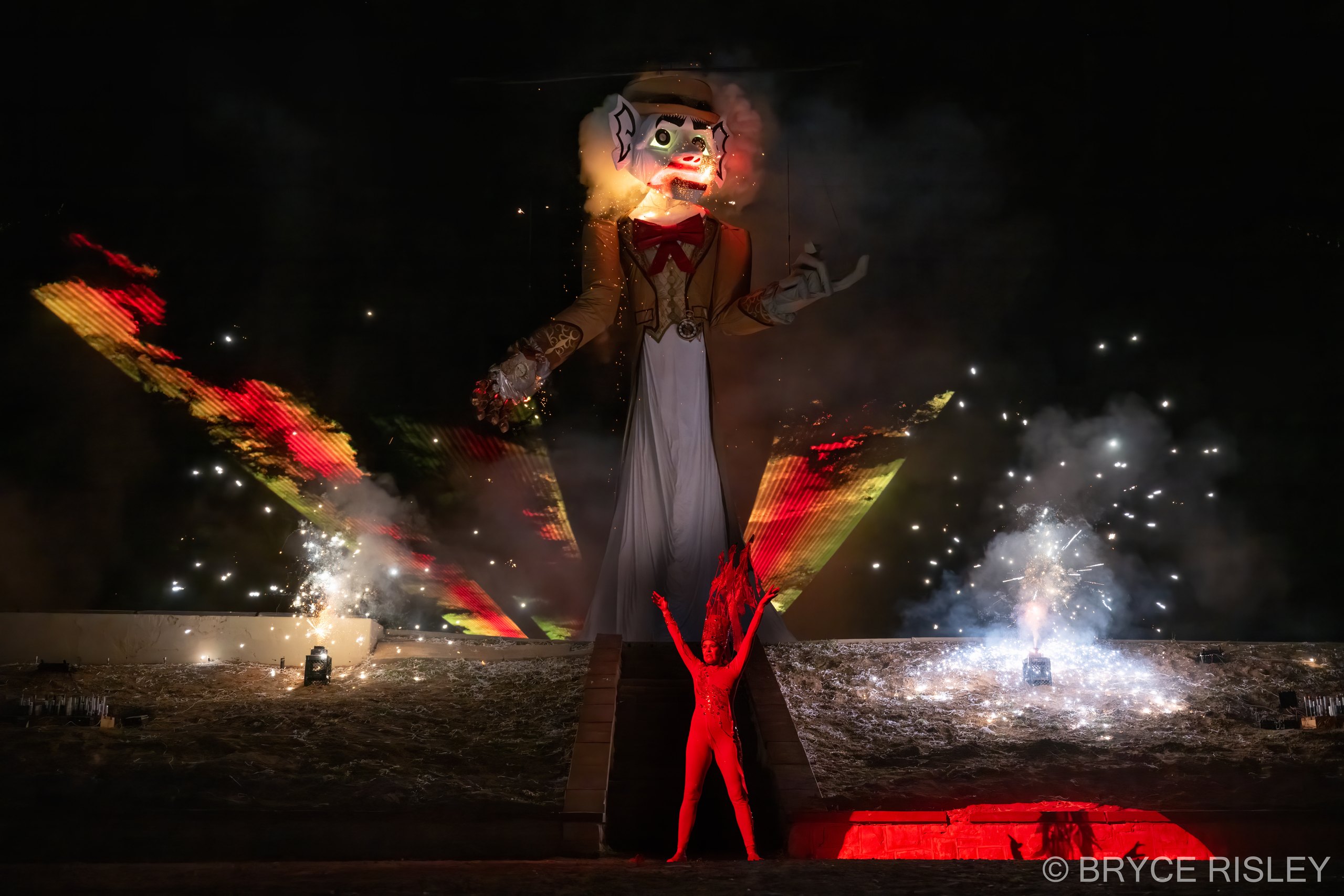 Zozobra, the fifty-foot marionette known as Old Man Gloom, alight at the head with arms outstretched over Fort Marcy Park at night, as the Fire Spirit in red raises both arms at the base of the platform and fireworks and sparks burst on either side.