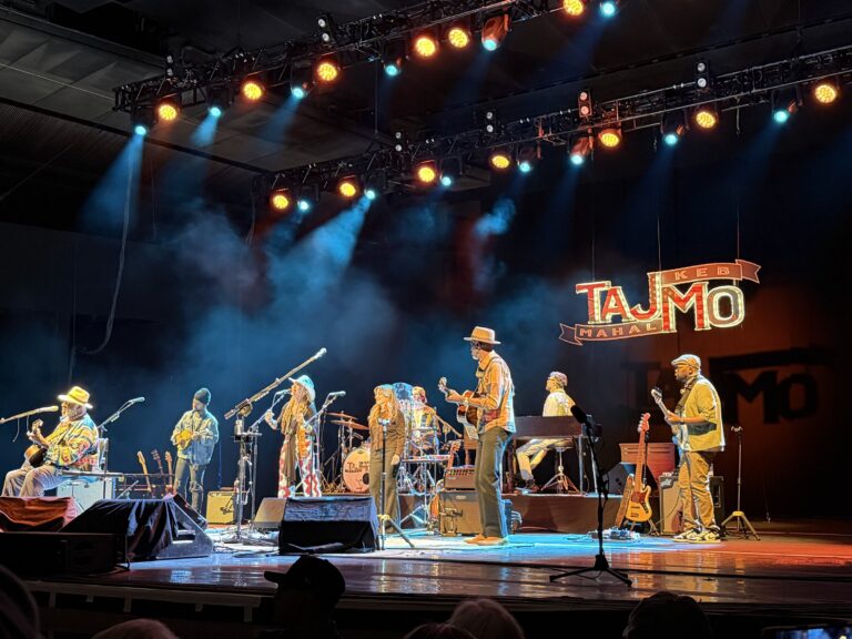Taj Mahal and Keb' Mo' on stage with their band at the Santa Fe Opera, both in straw hats with guitars, beneath a lit TajMo sign and orange and blue stage lights.