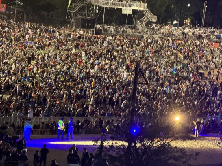 The crowd filling the field at Fort Marcy Park before the burning of Zozobra, seen from the hillside above, with bleachers and stage scaffolding beyond.